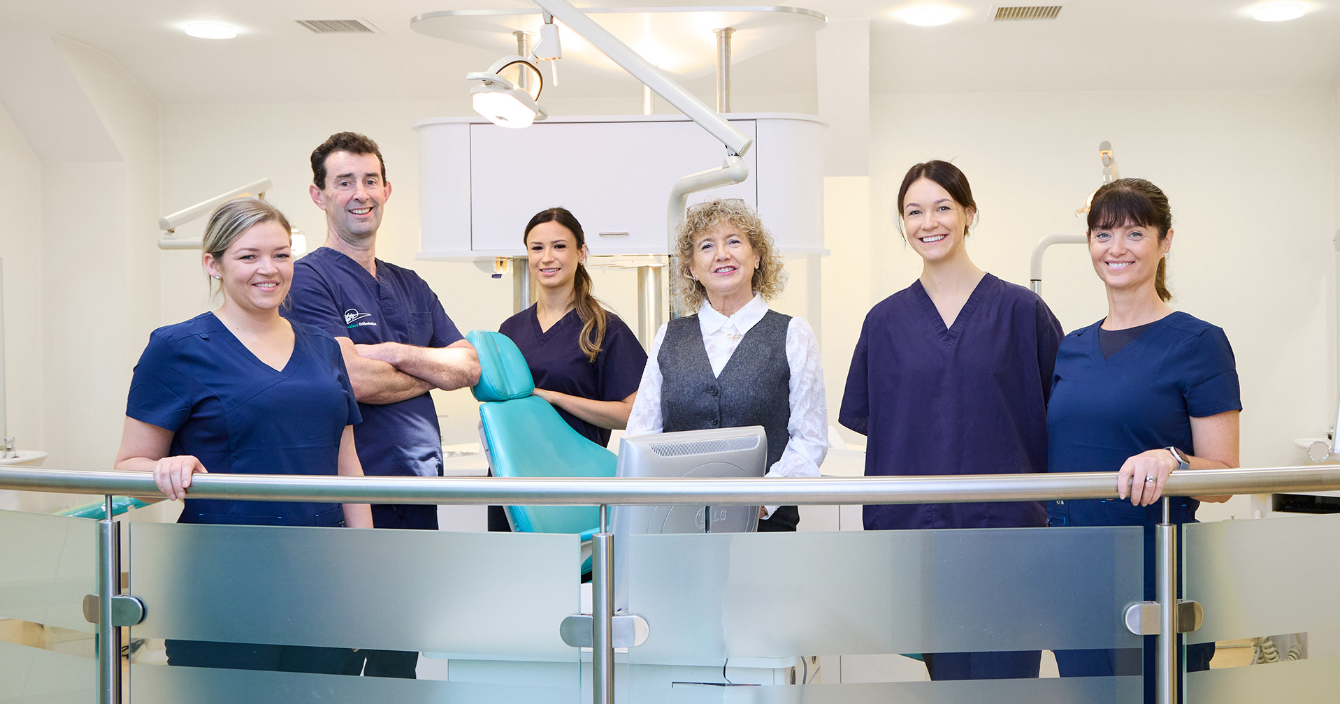 A team of six from Wexford Orthodontics, including dentists, dental assistants, and a receptionist, standing together in a modern dental clinic. They are dressed in navy blue scrubs and a white blouse with a vest, smiling confidently. The background features dental chairs, lights, and equipment