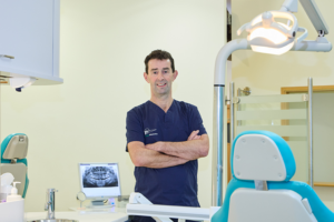 Picture of an Orthodontist in navy scrubs standing confidently in a modern dental clinic with arms crossed, a dental chair, and an X-ray image displayed on a computer screen in the background