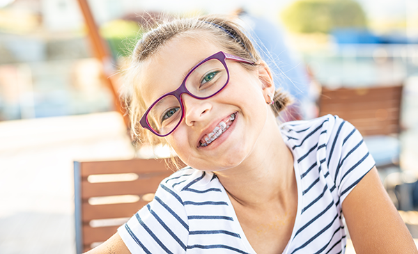 Picture of a young girl with light brown hair, wearing purple glasses and braces, smiles brightly while sitting outdoors at a wooden table. She is dressed in a white and navy striped shirt, and the background is blurred, showing an outdoor setting with warm sunlight