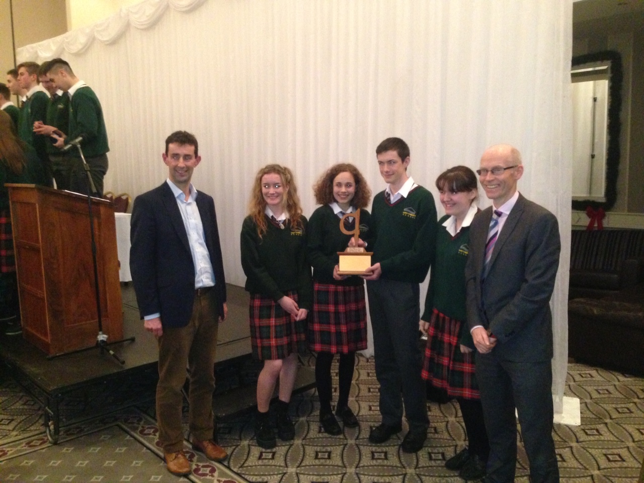 Picture of a Group of students in green sweaters and plaid skirts or pants posing with a trophy, alongside two men in formal attire, at an award ceremony on a stage with a white curtain backdrop