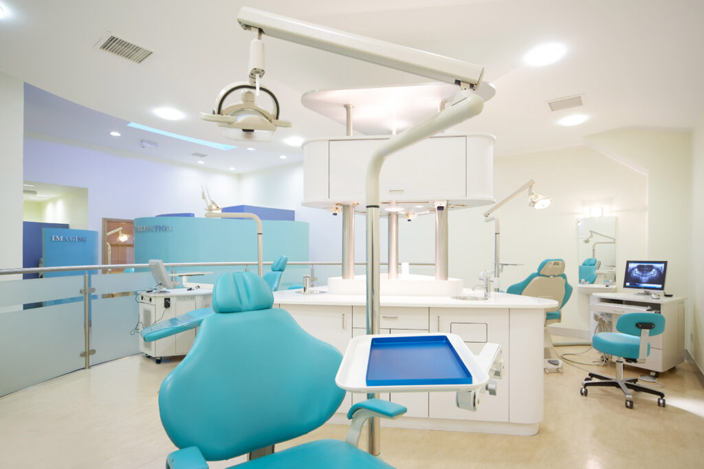 Bright and modern orthodontic clinic featuring multiple blue dental chairs, overhead examination lights, white cabinetry, and a computer displaying an orthodontic X-ray