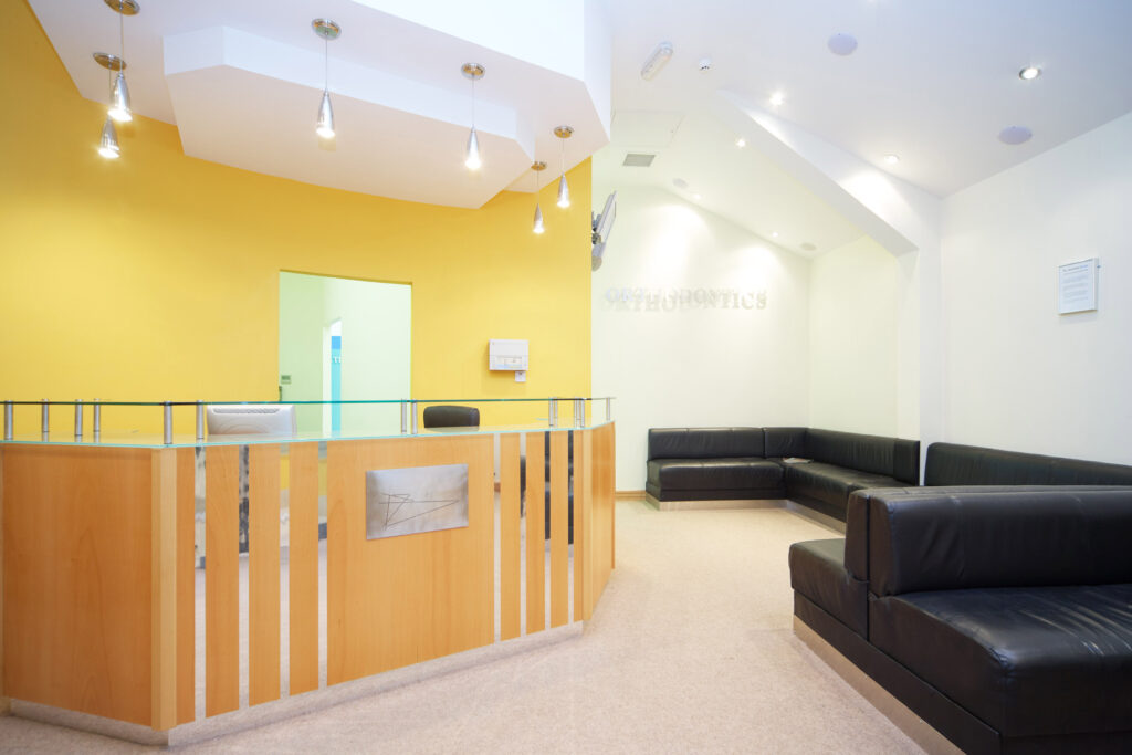 Modern dental clinic reception area with a wooden and glass reception desk, yellow accent wall, black leather seating, and bright ceiling lights