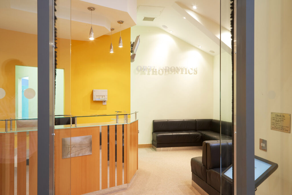 Modern orthodontic office reception area featuring a wooden reception desk with glass counter, bright yellow accent wall, comfortable black leather seating in the waiting area, and subtle 'ORTHODONTICS' signage in a clean, professional environment"