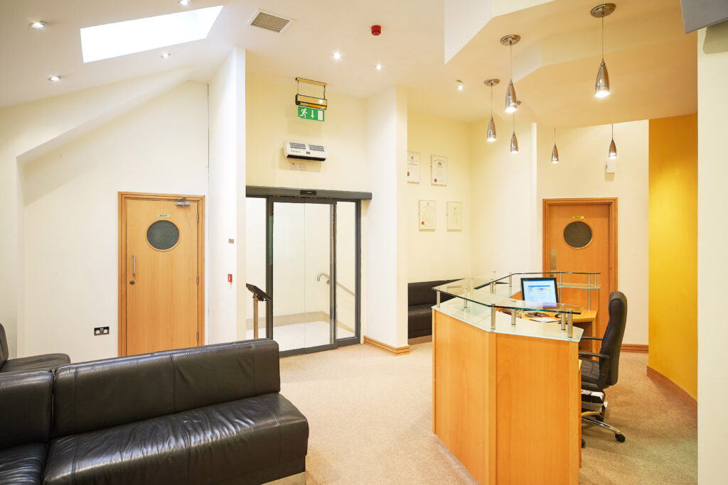 "Orthodontic office reception area with wooden reception desk, computer workstation, black leather waiting area seating, automatic sliding glass entrance doors, wooden treatment room doors, high ceilings with skylights, and modern pendant lighting. The bright, contemporary space features cream walls with a yellow accent wall"