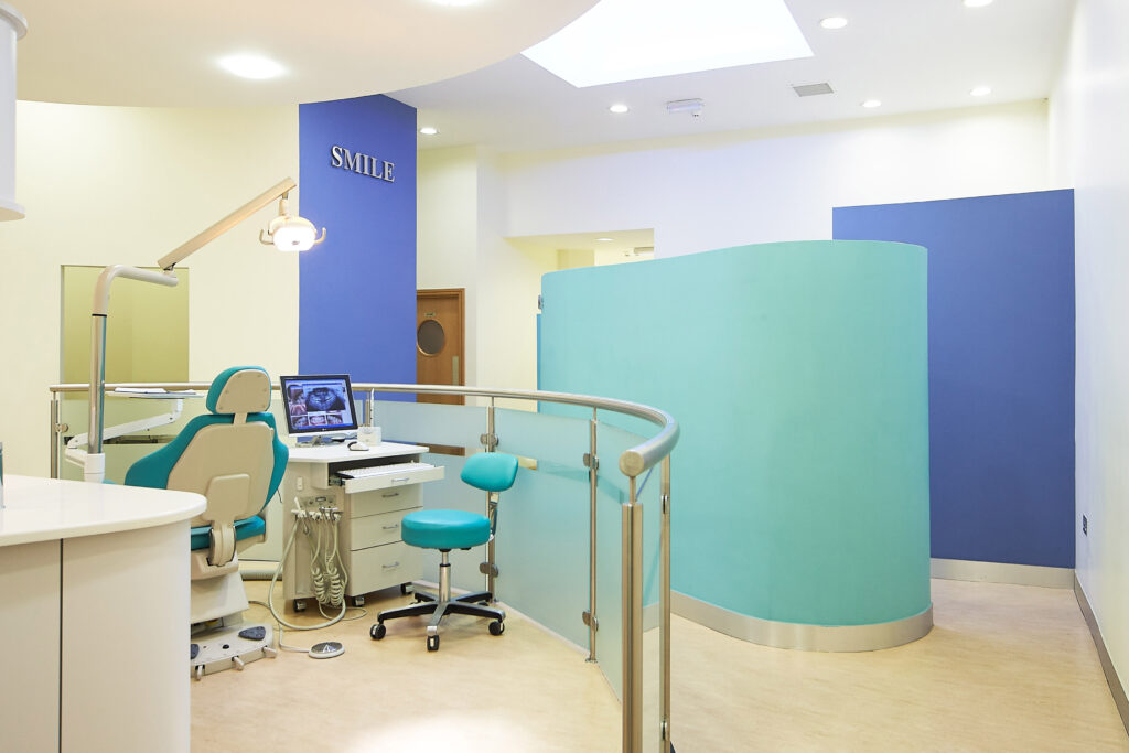 "Modern orthodontic treatment bay featuring a teal dental chair, dentist's stool, and computer monitor displaying dental x-rays. The space has a contemporary design with curve