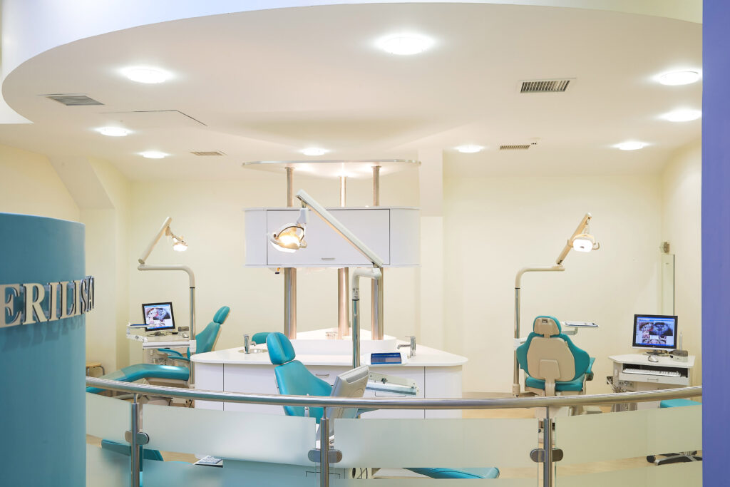 "Open-concept orthodontic treatment area with multiple teal dental chairs arranged around a central workstation. Each chair has its own dental light and computer monitor displaying dental images. The modern space features sleek cabinetry, glass privacy panels, curved ceilings with recessed lighting, and blue accent walls with 'BRACES' lettering visible. This contemporary clinical environment is designed for efficient orthodontic treatment"