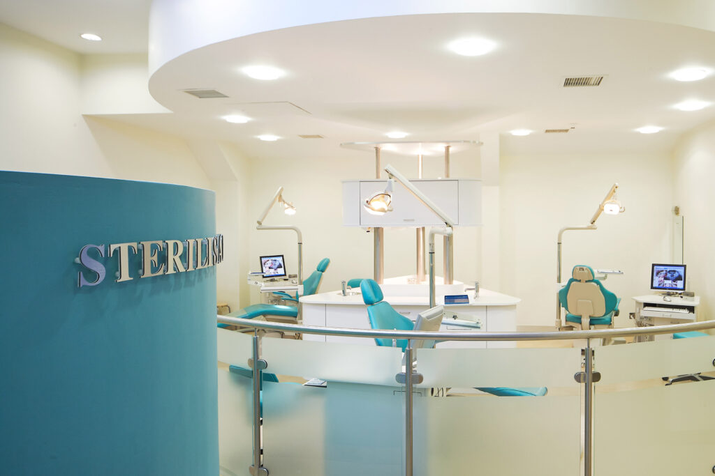 "Open-concept orthodontic treatment area with multiple teal dental chairs arranged around a central workstation. Each chair has its own dental light and computer monitor displaying dental images. The modern space features sleek cabinetry, glass privacy panels, curved ceilings with recessed lighting, and blue accent walls with 'BRACES' lettering visible. This contemporary clinical environment is designed for efficient orthodontic treatment"