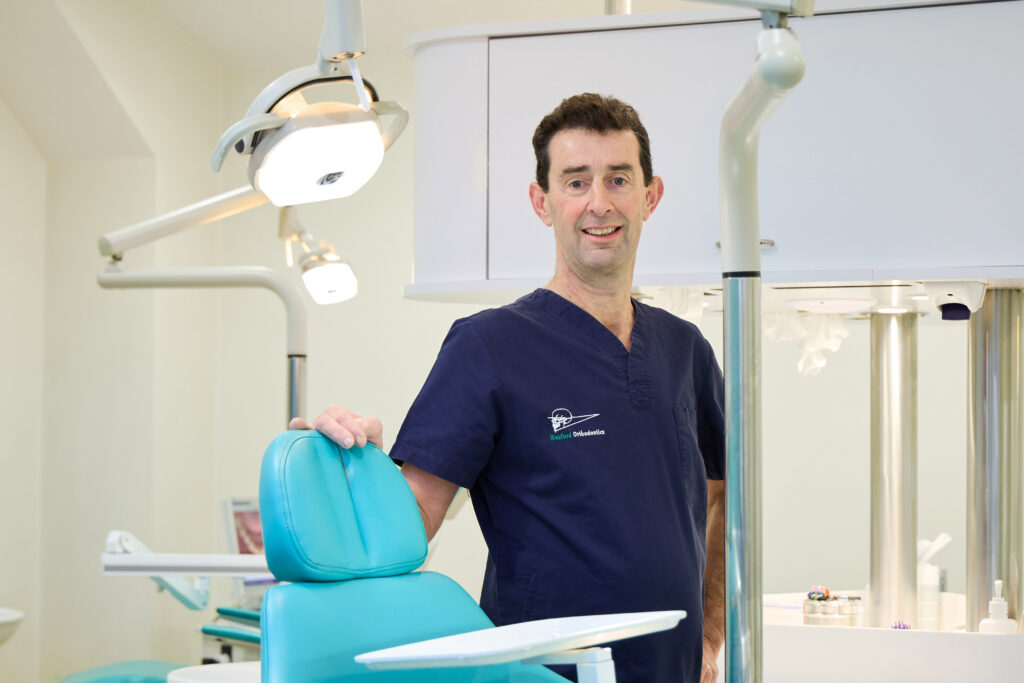 Picture of an Orthodontist in navy scrubs standing confidently in a modern dental clinic with his hands on a teal dental chair and a slight smile