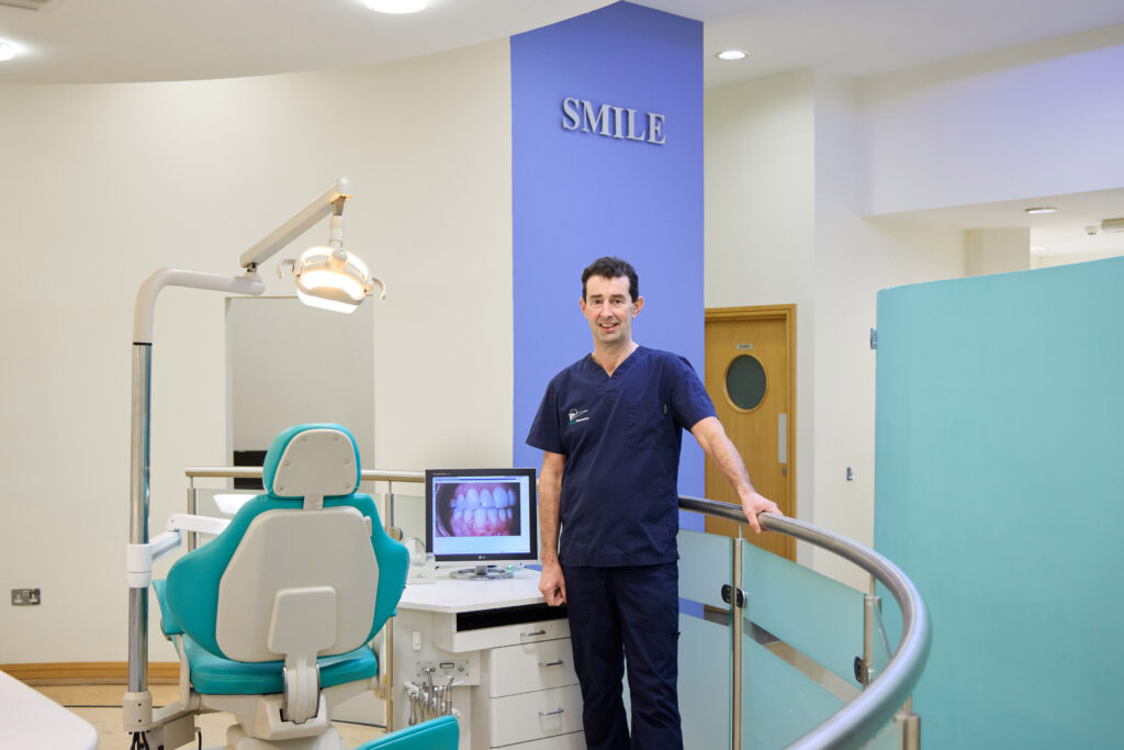 zoomed out Picture of an Orthodontist in navy scrubs in an open spaced orthodontic room with a computer screen with an image of teeth a teal dental chair and a blue wall with the text "SMILE"