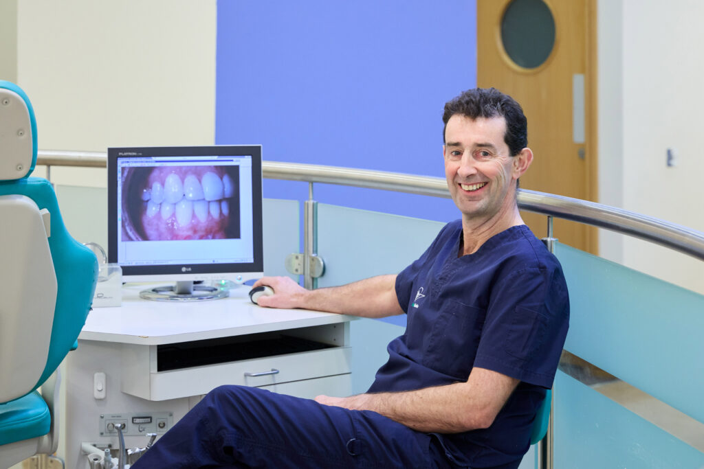 Picture of an Orthodontist in navy scrubs in an open spaced orthodontic room with a computer screen with an image of teeth