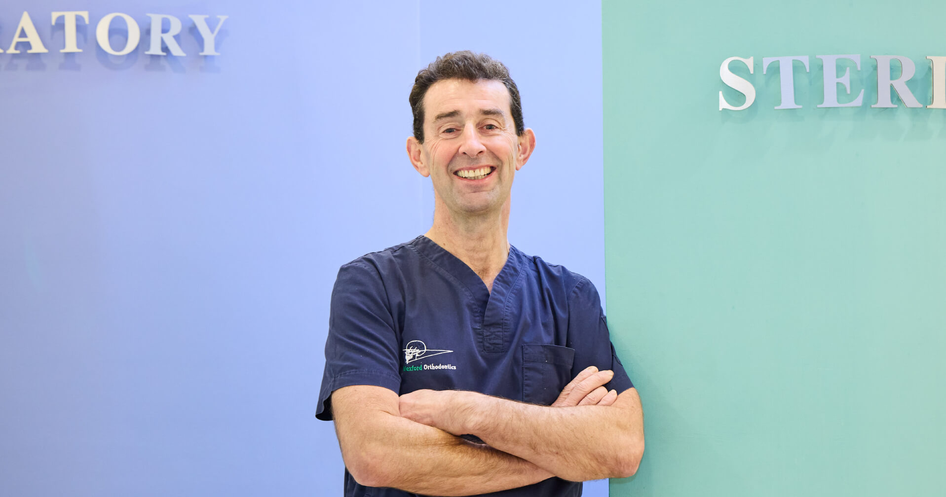 picture of a Male orthodontist in navy scrubs standing with arms crossed, smiling in a clinical setting with 'STER' and 'ATORY' visible on the wall behind him