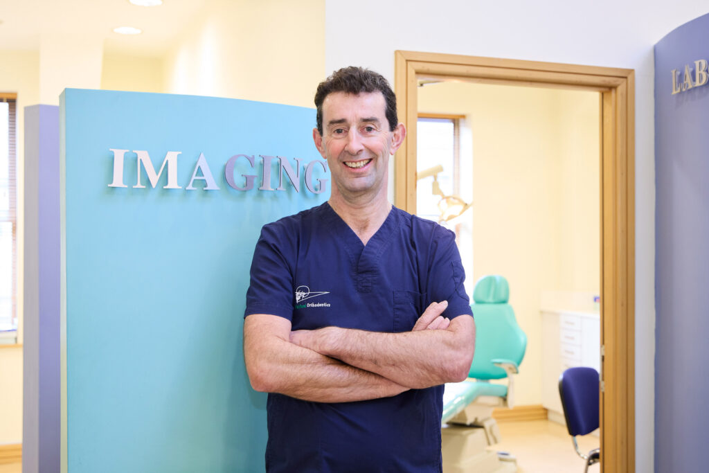 Picture of an Orthodontist in navy scrubs standing infront of a teal wall with the signage imaging and a room with a teal dental chair in the backround