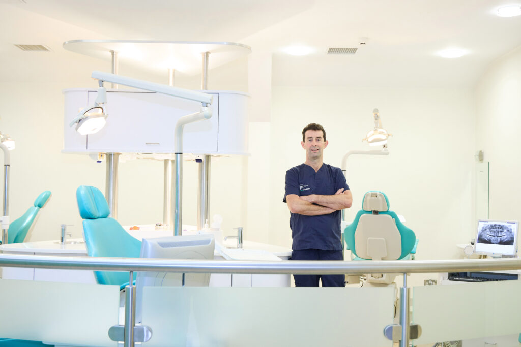 Picture of an Orthodontist in navy scrubs standing in an open spaced orthodontics room with multiple teal dental chairs orthodontic lights and a computer screen with an x-ray of a jaw