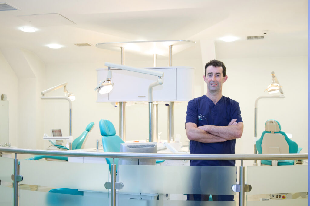 Zoomed in Picture of an Orthodontist in navy scrubs standing in an open spaced orthodontics room with multiple teal dental chairs orthodontic lights and a computer screen with an x-ray of a jaw