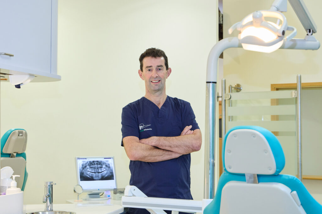 Picture of an Orthodontist in navy scrubs standing in an orthodontics room with a teal dental chair an orthodontic light and a computer screen with an x-ray of a jaw