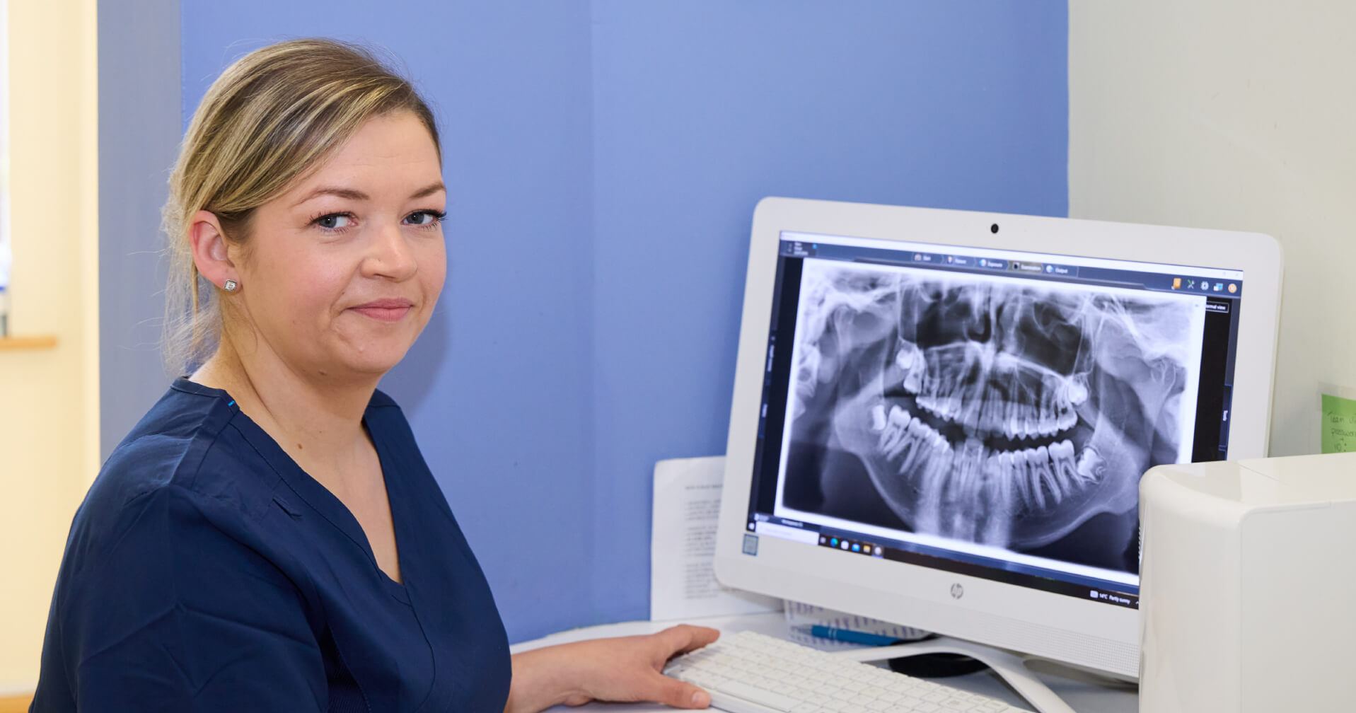 Female orthodontics professional in navy scrubs sitting at a desk with a computer displaying a panoramic orthodontic X-ray