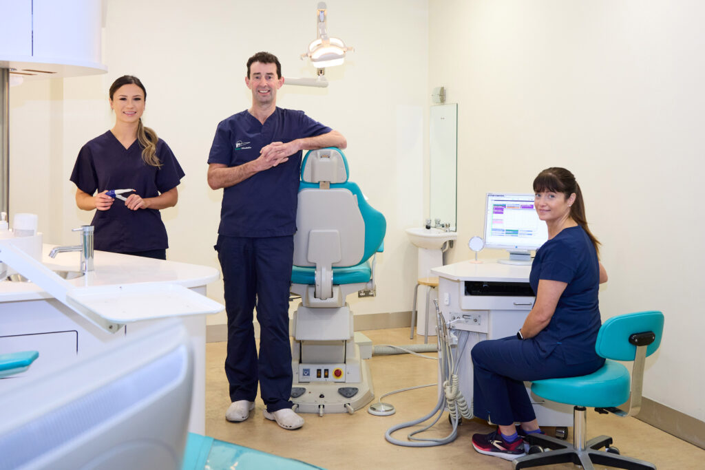 Picture of an orthodontist and two orthodontic assistants in navy scrubs in a large orthodontic room smiling at the camera