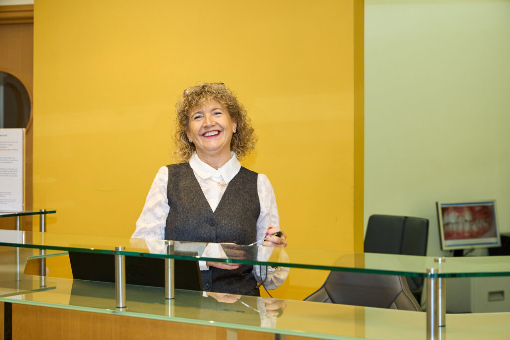 picture of A smiling woman with curly blonde hair standing behind a glass reception desk, wearing a white blouse with a dark gray vest, against a yellow wall. A dental office setting is visible in the background, with a computer screen displaying dental images