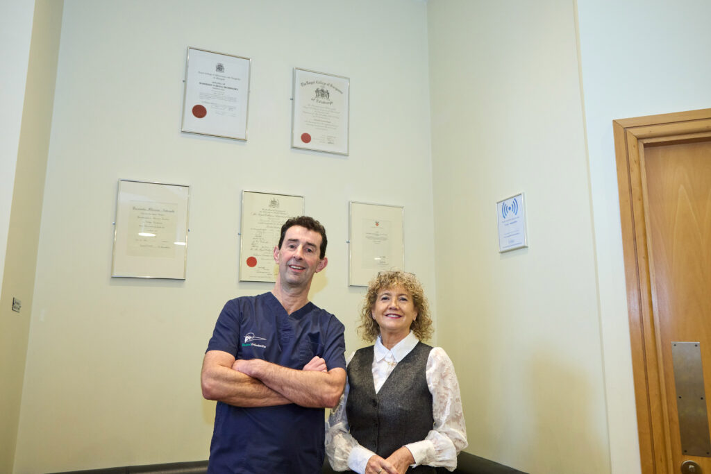 picture of an orthodontist in navy scrubs and a receptionist standing infront of a wall that has certifications and awards hung up on it