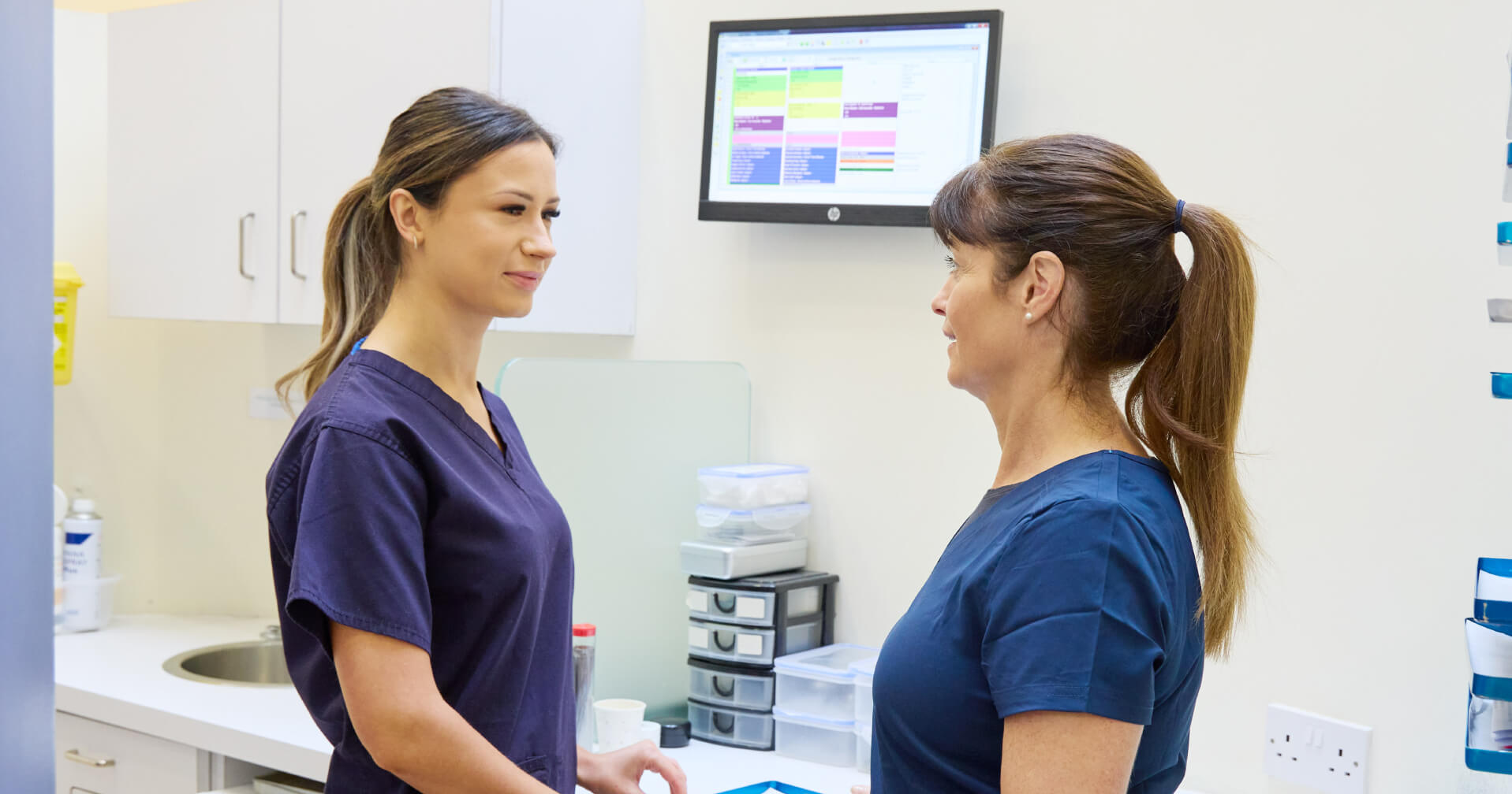 picture of Two female orthodontics professionals in scrubs discussing in a clinical setting with a digital schedule displayed on the wall.