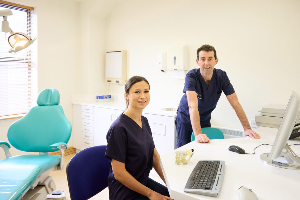 picture of a two orthodontists in navy scrubs in an orthodontic room with a computer on a desk and a teal dental chair with a light above it