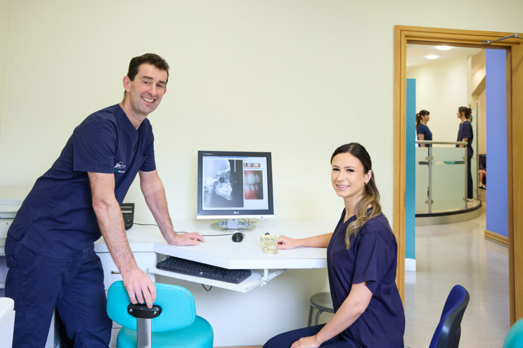 picture of a two orthodontists in navy scrubs in an orthodontic room with a computer on a desk with an x-ray and two more orthodontists in the background