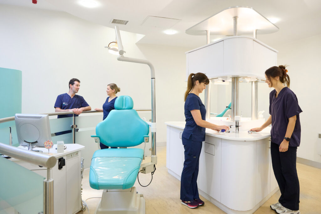 picture of a four orthodontists in navy scrubs in an orthodontic room with a computer on a desk and a teal dental chair with a light above it and a large island counter in the middle