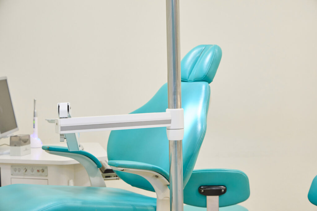 A close-up of a teal dental chair with a metal pole and arm, and a white countertop with equipment in the background