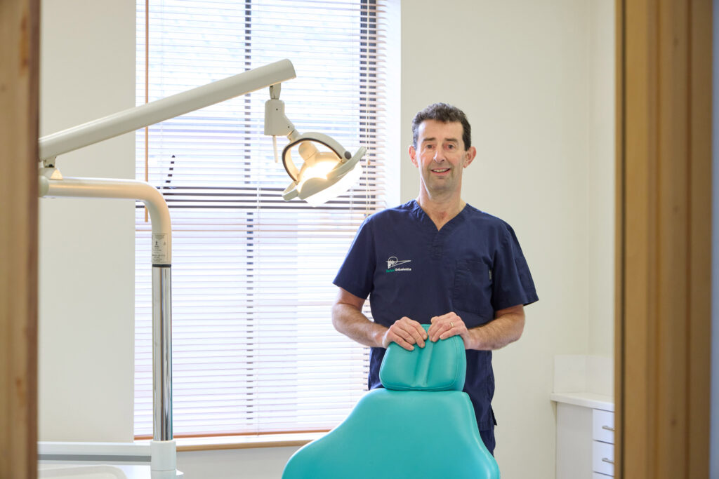 picture of an orthodontist in navy scrubs stood behind a teal dental chair with a light above it and a window and blinds in the background